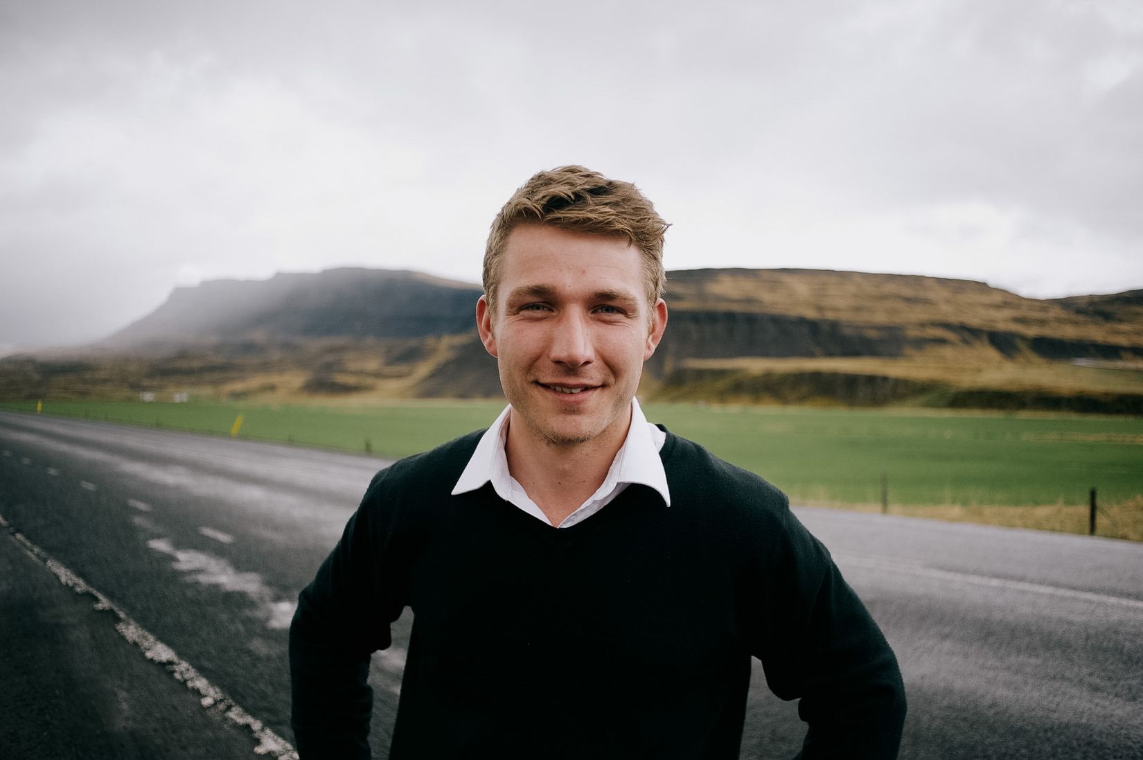 A fair-haired man in a dark jumper and white collar standing in the middle of an empty road, hands on hips