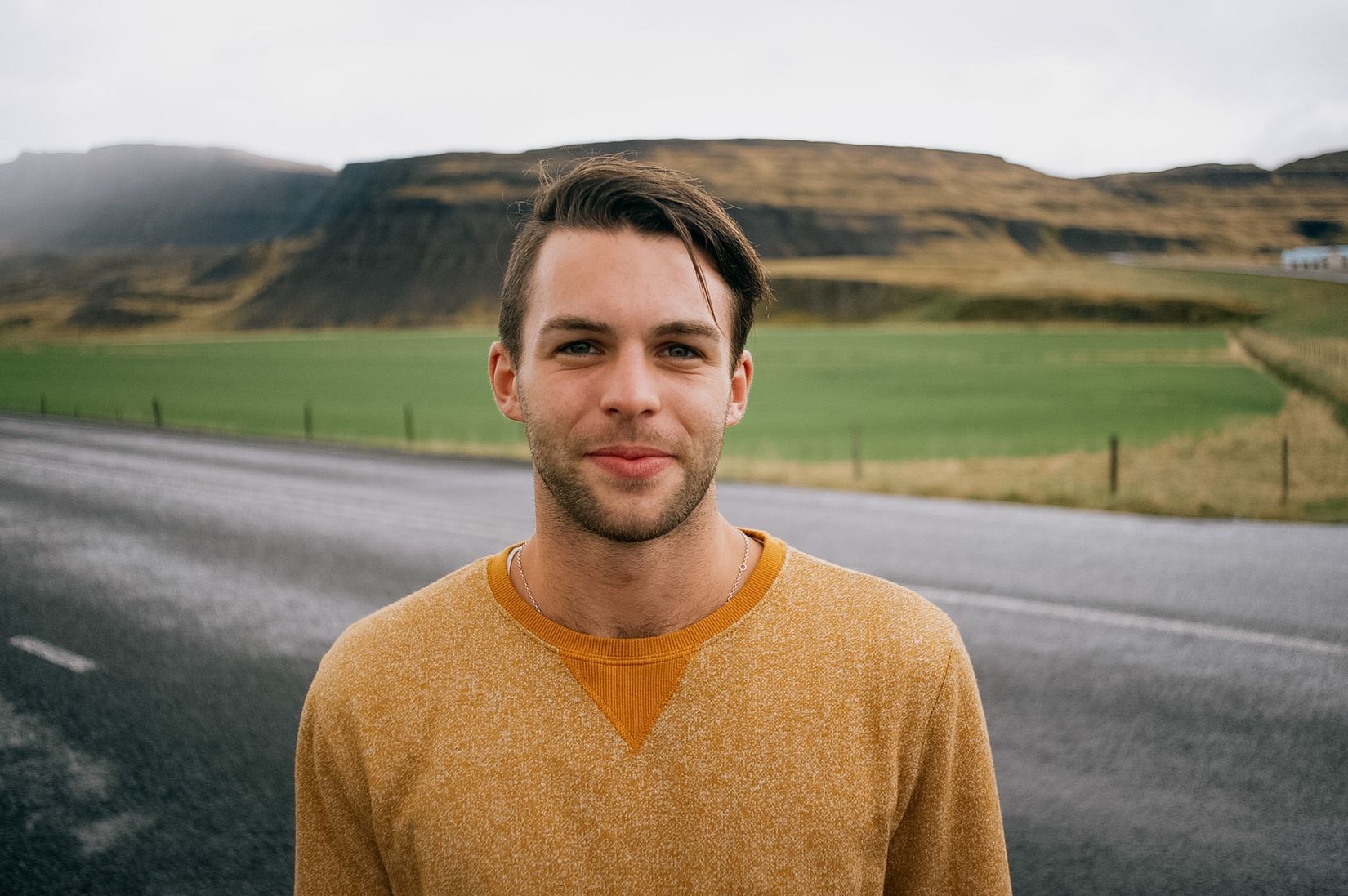 A dark-haired man in a mustard jumper on the same road, green field and a flat-topped hill behind him