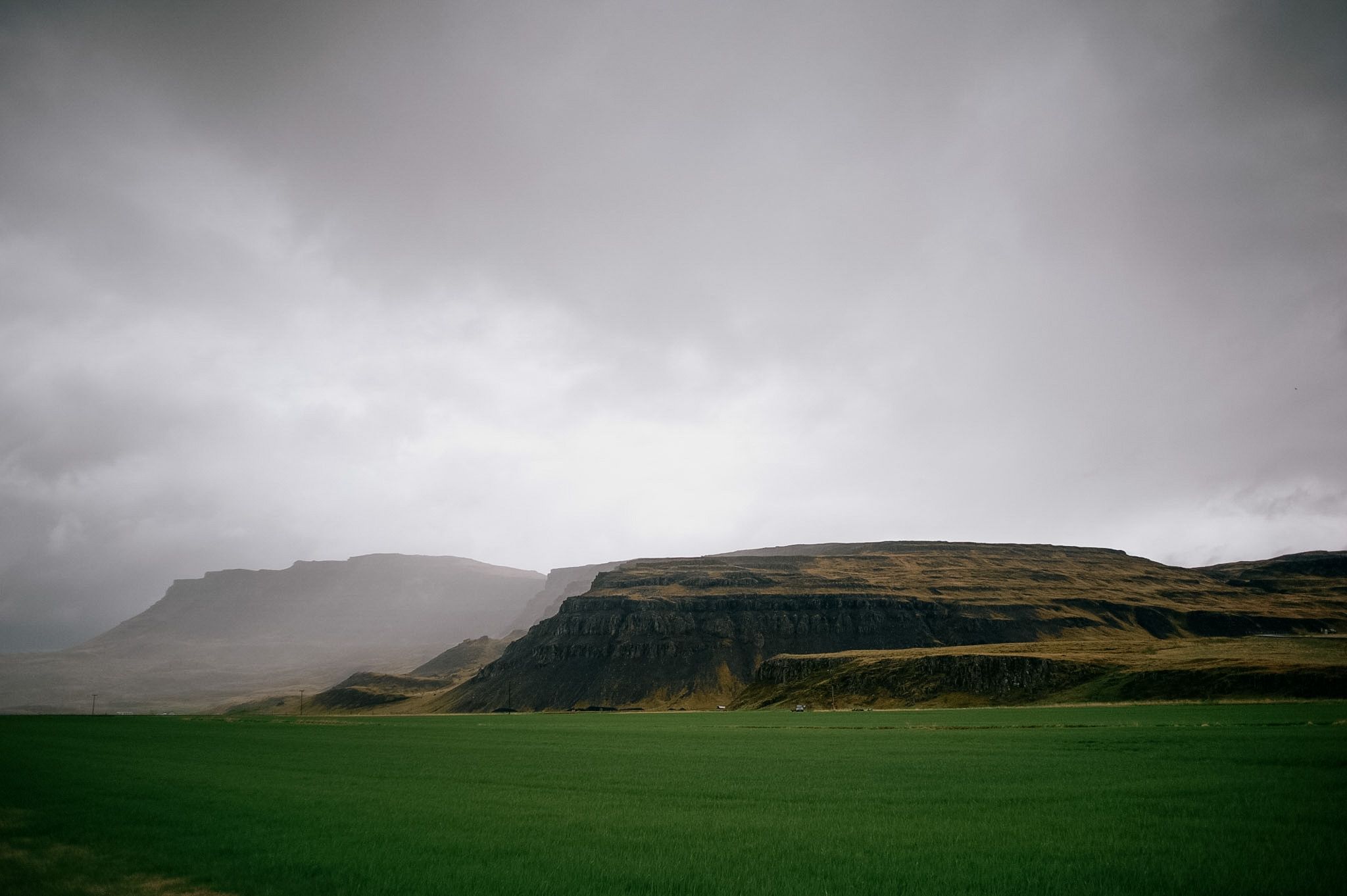 A flat-topped escarpment above a bright green field, the whole sky one sheet of rain-grey cloud