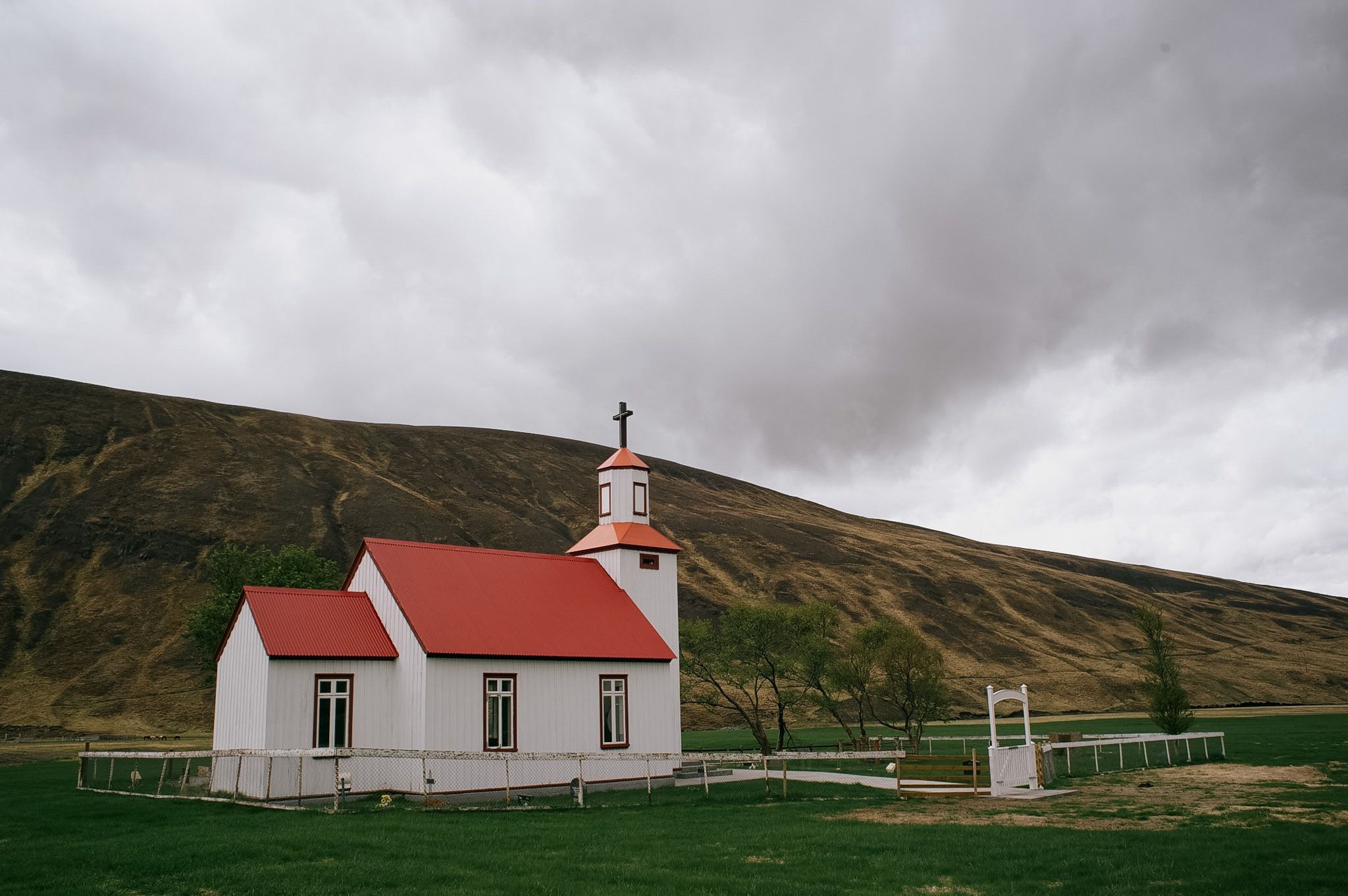 A small white church with a red roof and steeple alone on the grass below a brown hill