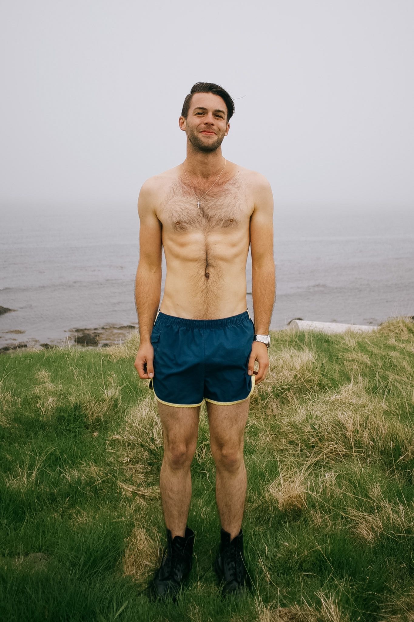 A man in blue shorts and walking boots standing in the long grass above the shore, sea and fog behind him
