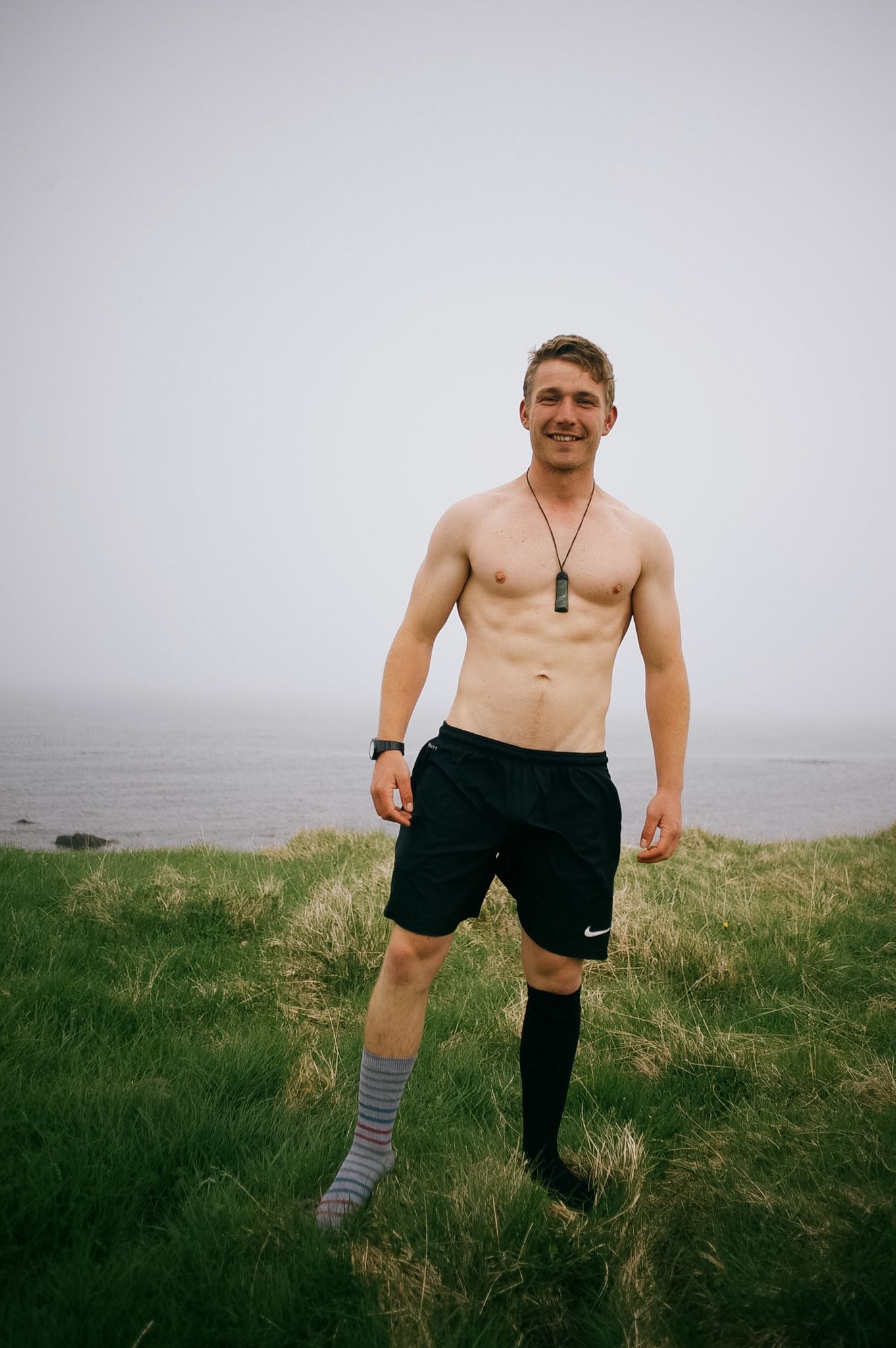 A man in black shorts and one striped sock grinning on the same bank, the water behind him white with fog