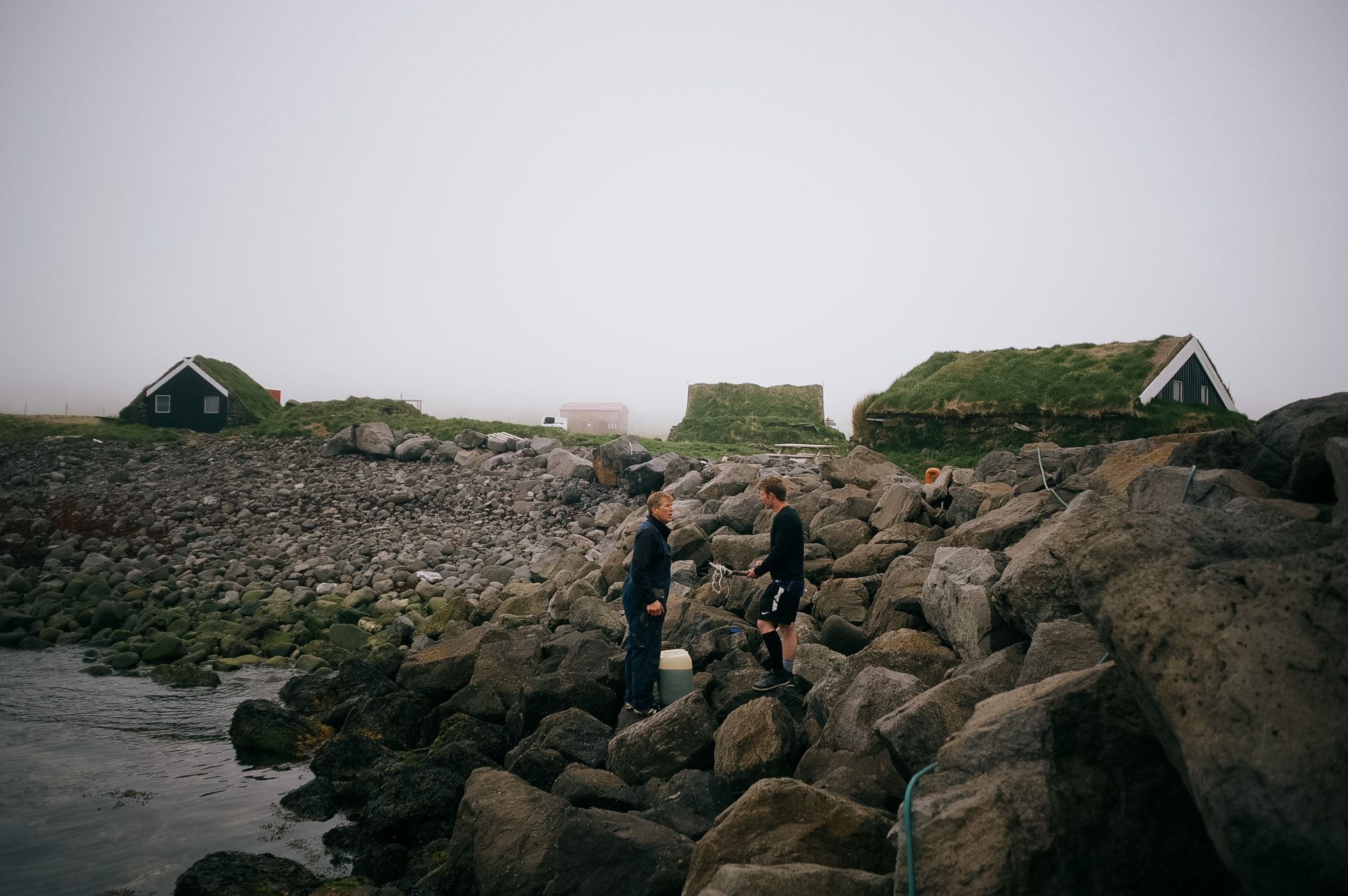 Two figures picking their way over wet boulders below a row of turf-roofed cottages in the mist