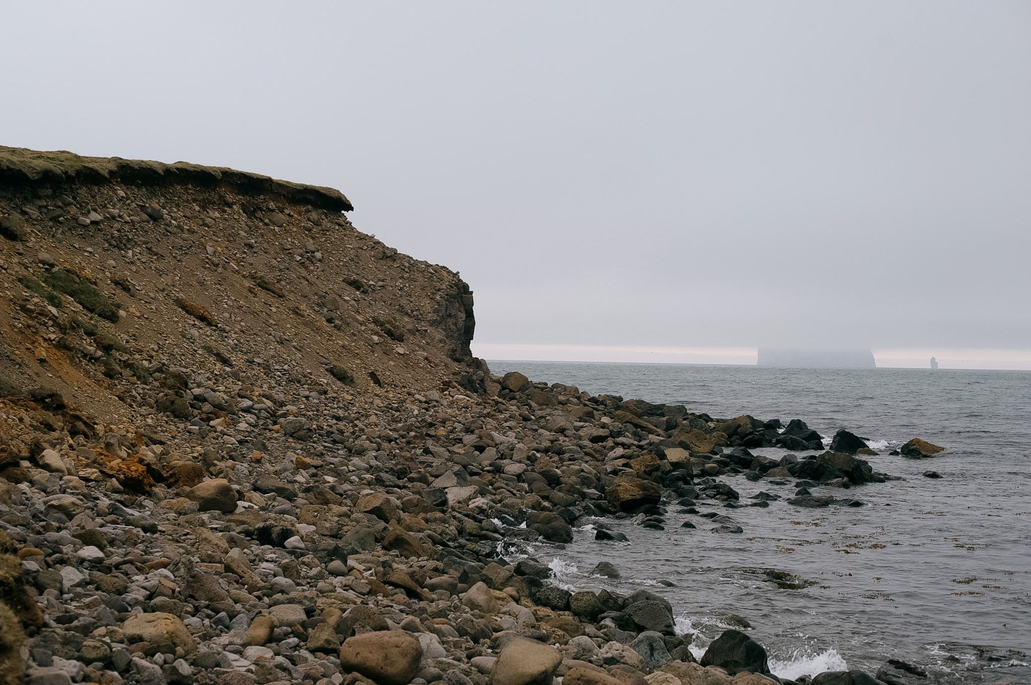 A crumbling earth cliff dropping to a stony beach, a flat-topped island just visible in the haze offshore