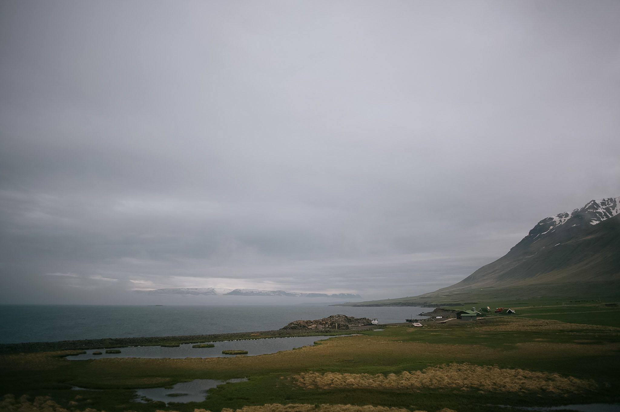 A farm and its green home field on a spit between the sea and a chain of pools, mountains fading up the fjord