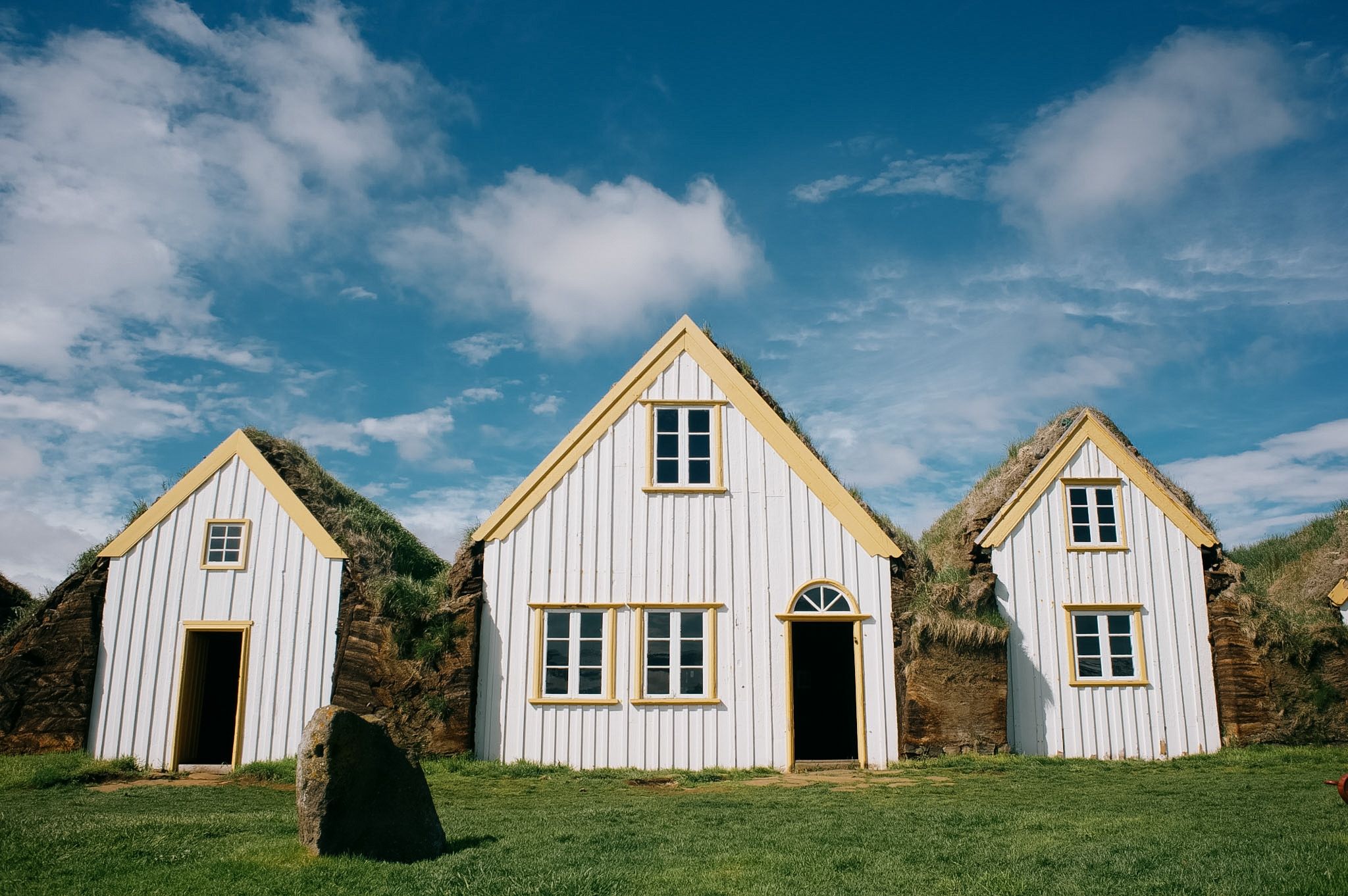 Three white gabled turf houses in a row, grass growing over their roofs and down their sides, under a hard blue sky