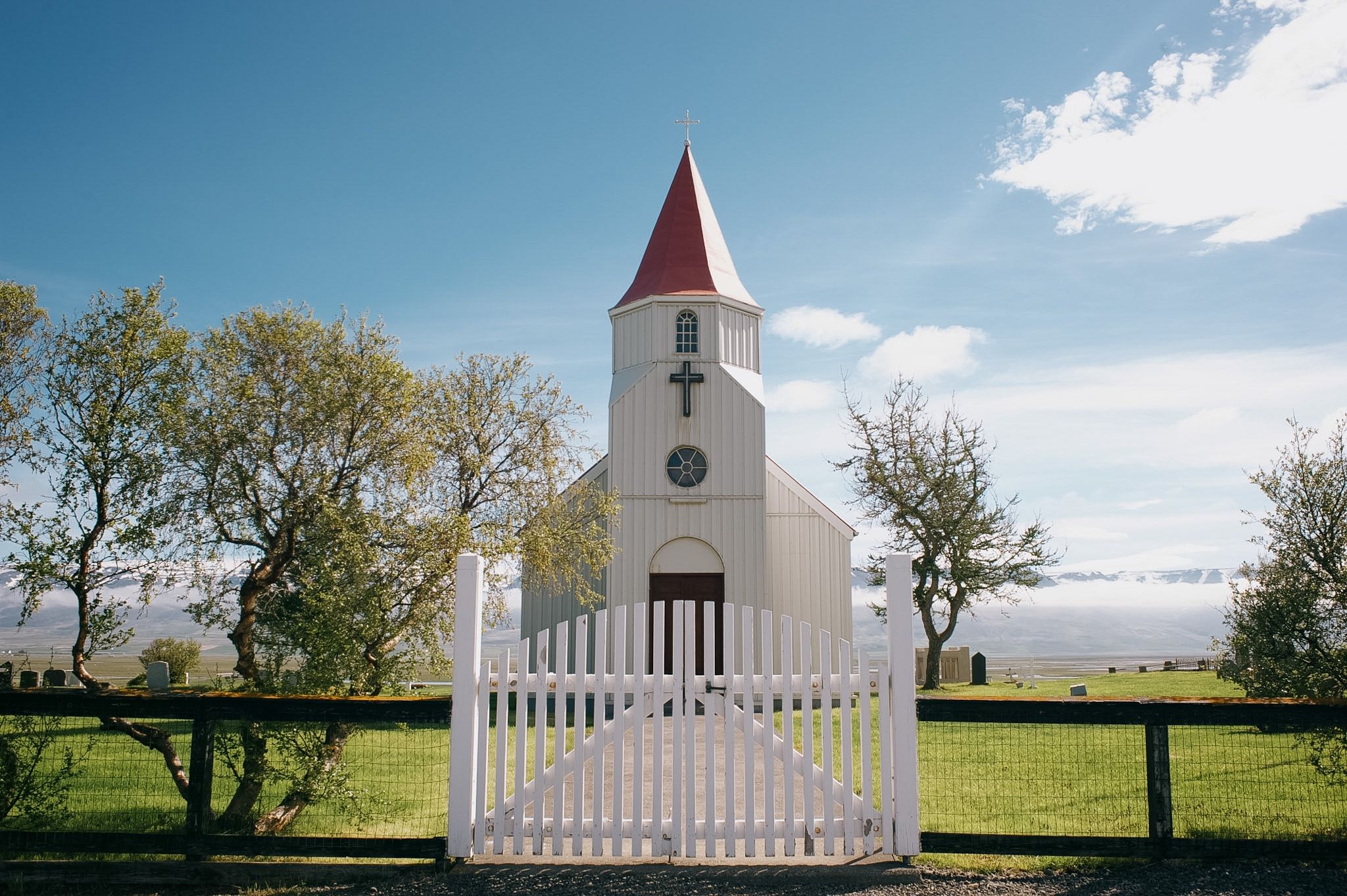 A white timber church with a red spire behind a picket gate, graves and bare trees either side