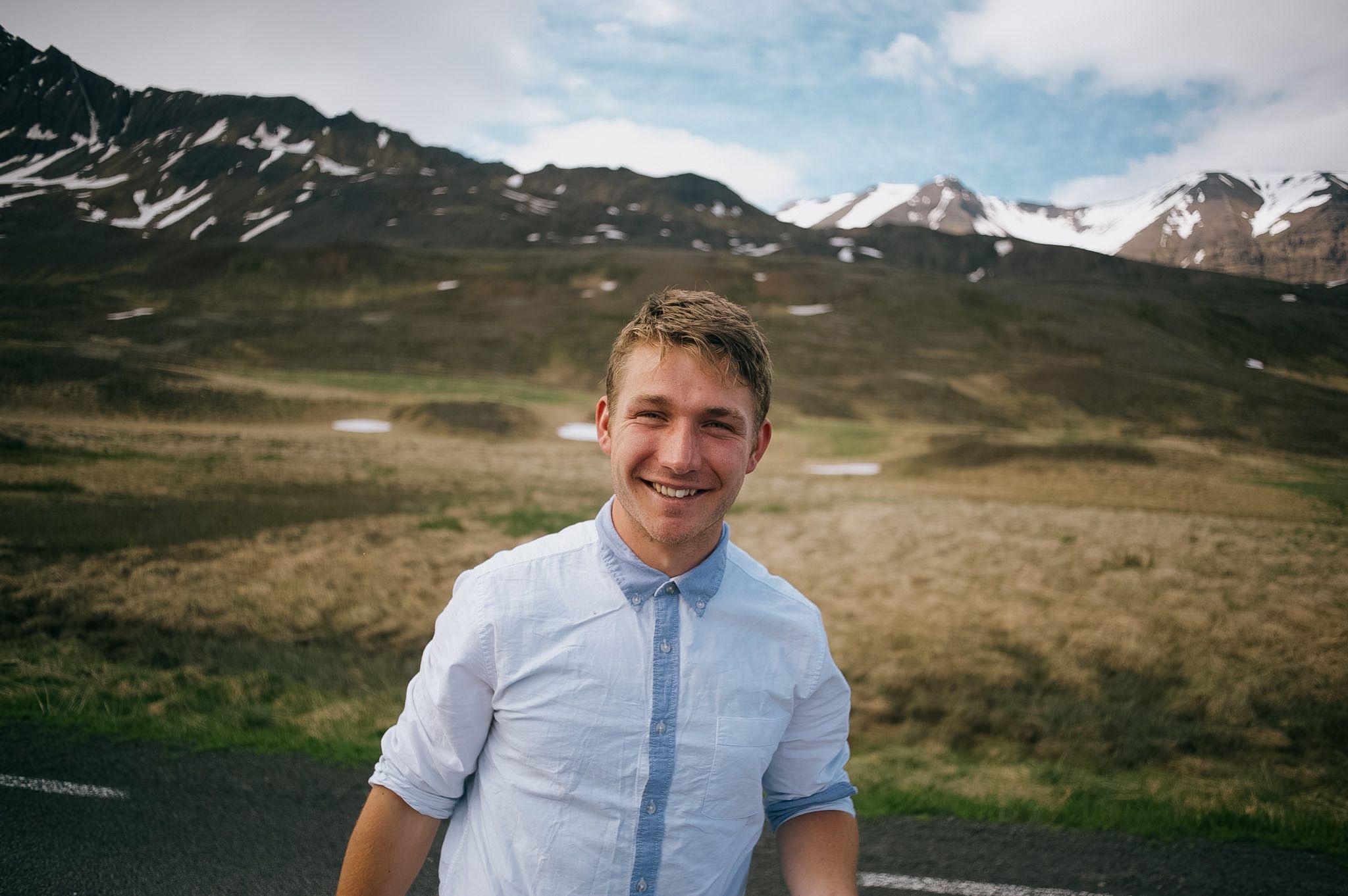 A fair-haired man in a pale blue shirt on the roadside, snow still lying in the gullies of the peaks behind