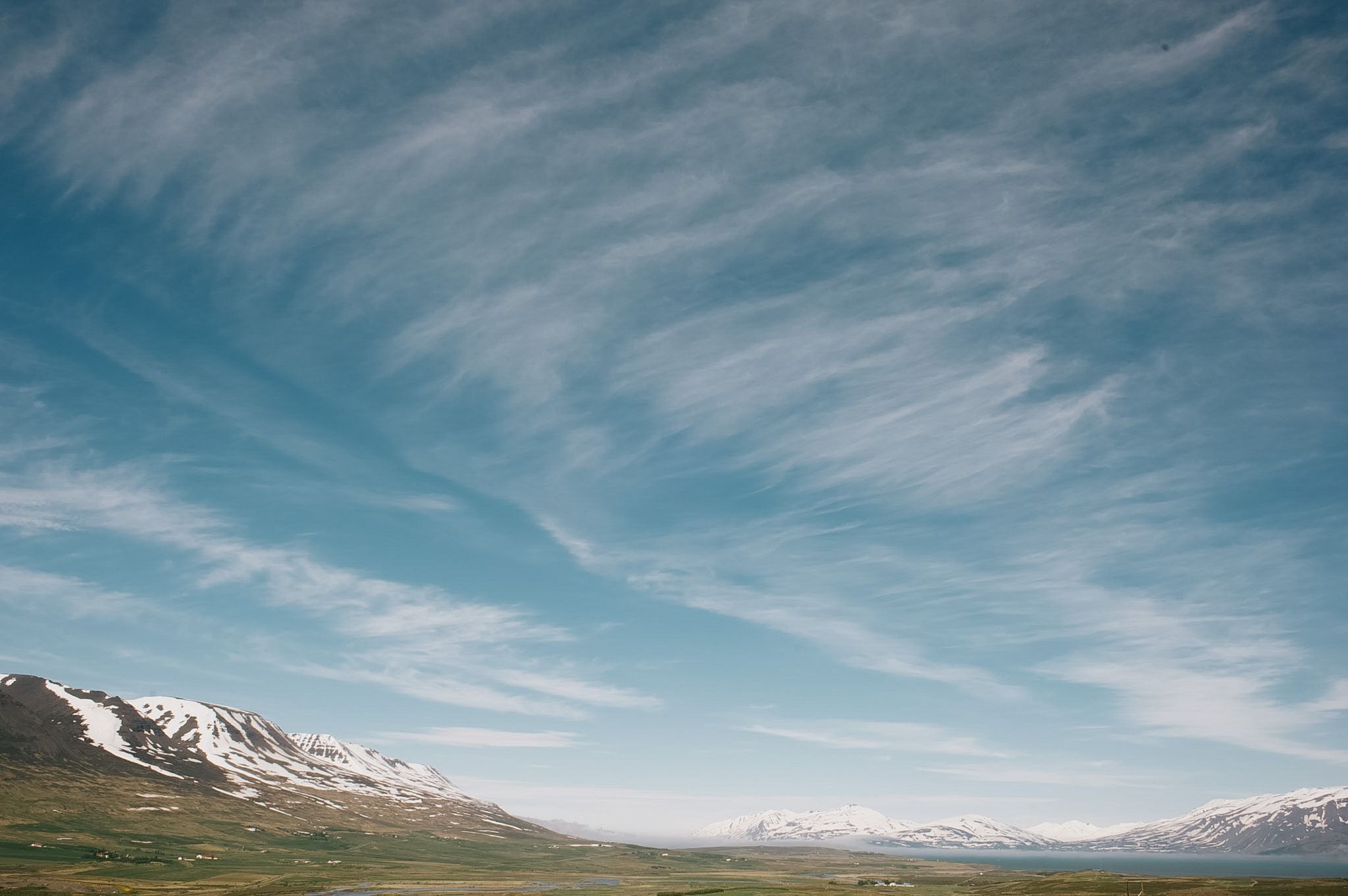 Cirrus drawn in long streaks across the sky above a snow-topped ridge and a flat green valley floor