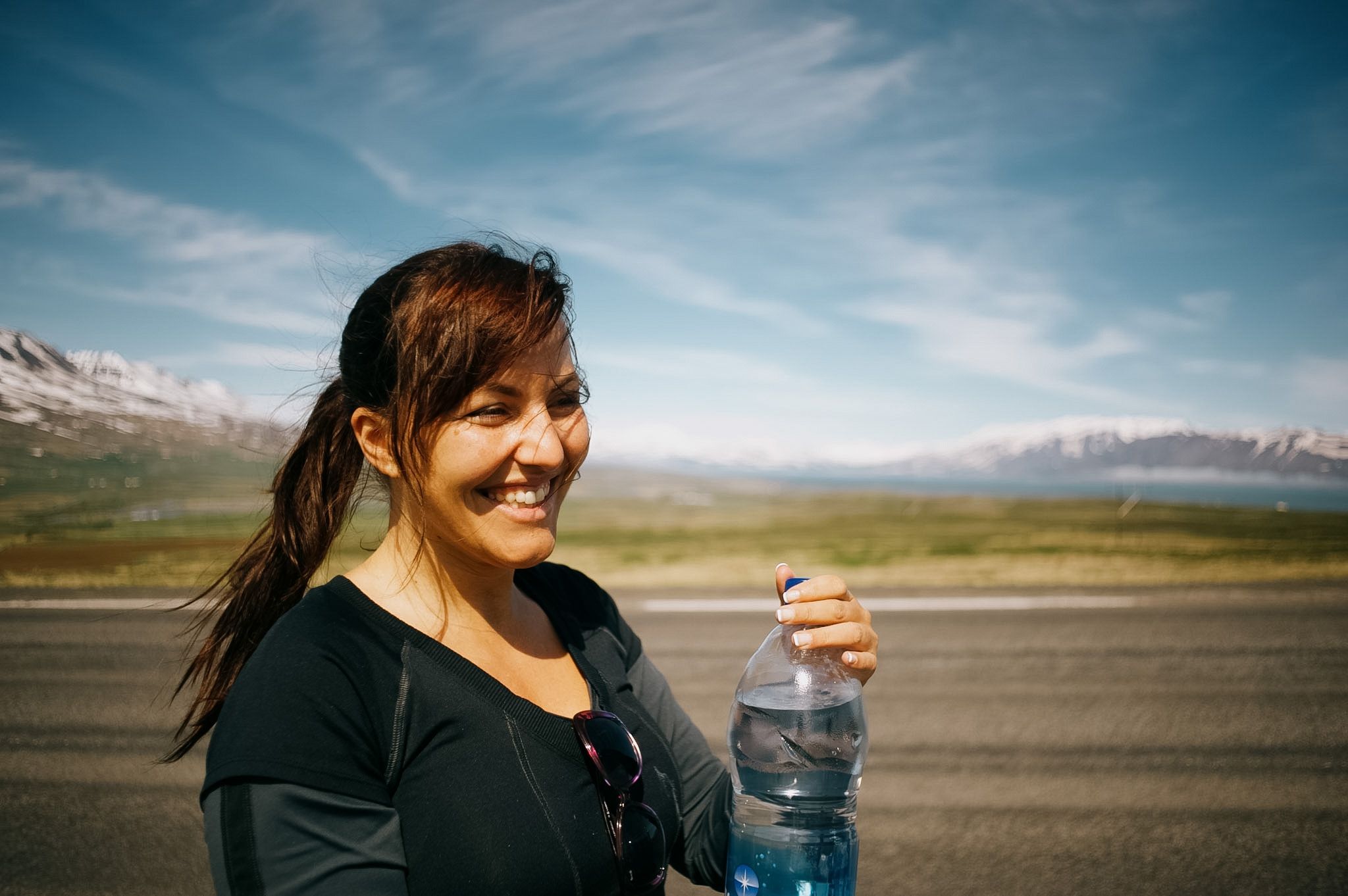 A woman laughing on the verge with a water bottle in her hand, sunglasses hooked on her collar, snow on the far shore