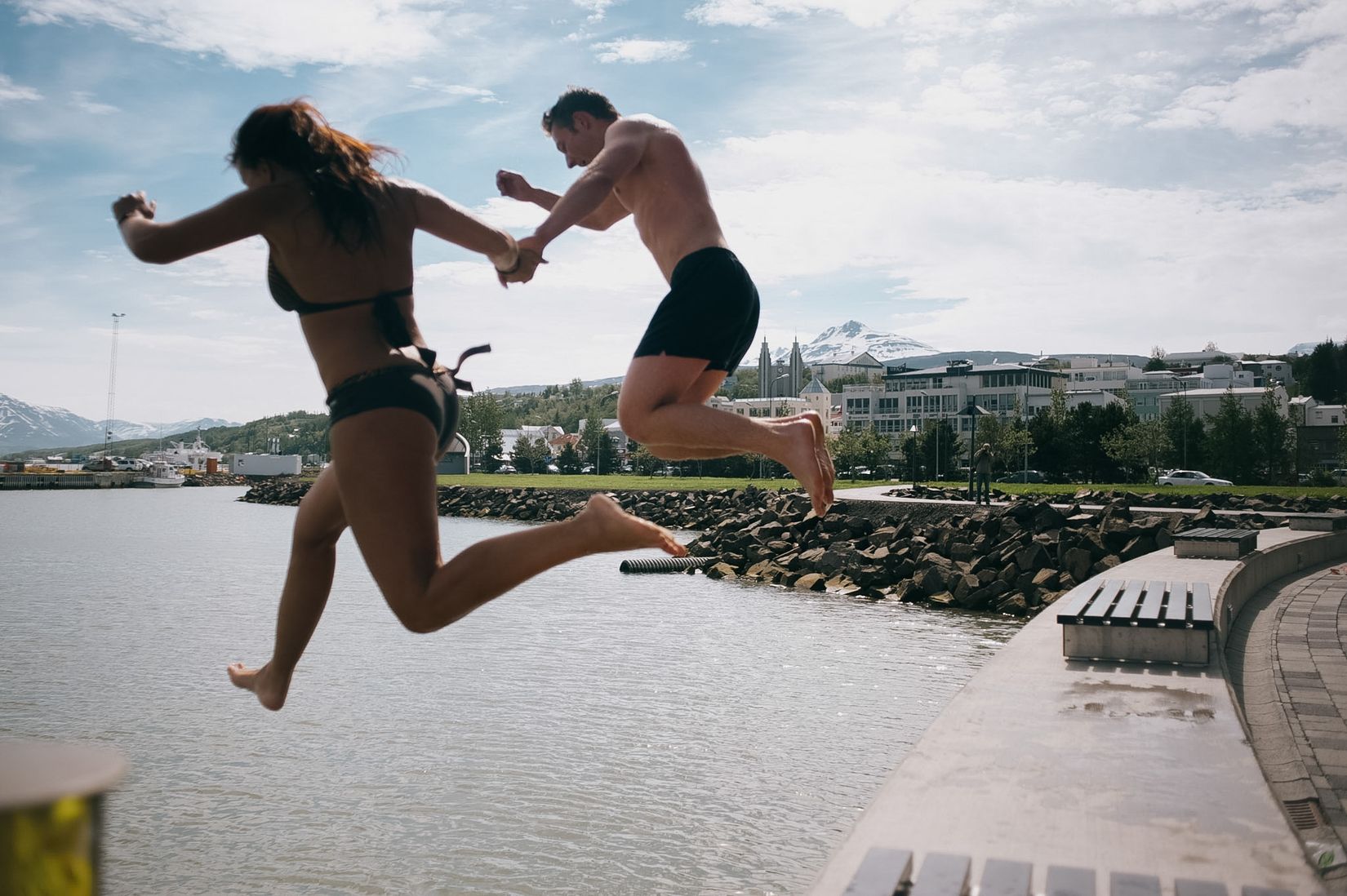 Two people jumping off a harbour wall hand in hand, mid-air over the water, the town and its mountains behind