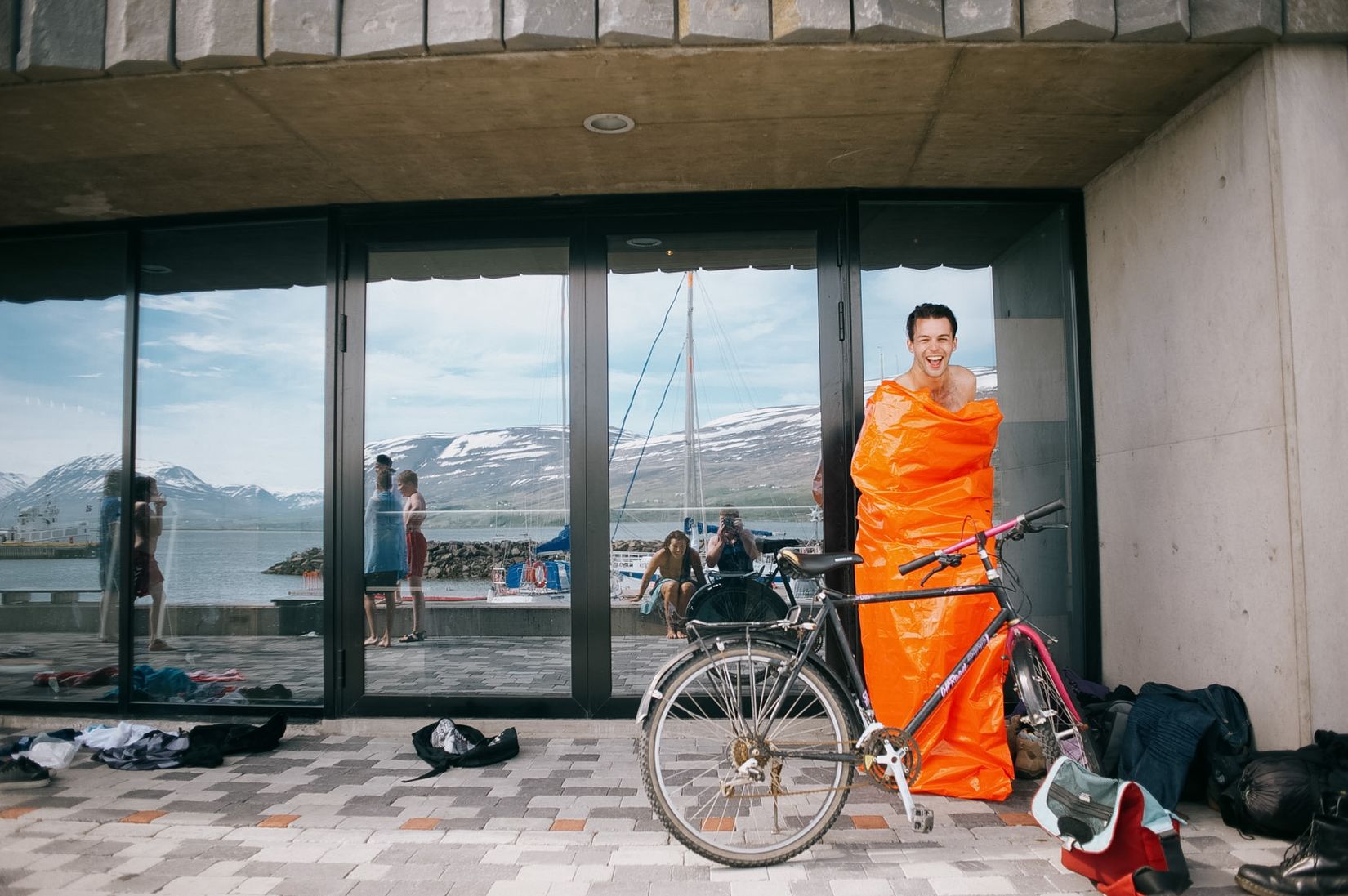 A man wrapped to the chin in an orange sleeping bag beside a bicycle, clothes strewn over the paving outside the pool