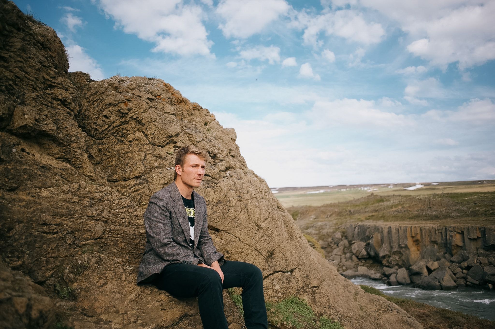 A man in a grey jacket sitting against a boulder above a basalt gorge, river running out across the plain below
