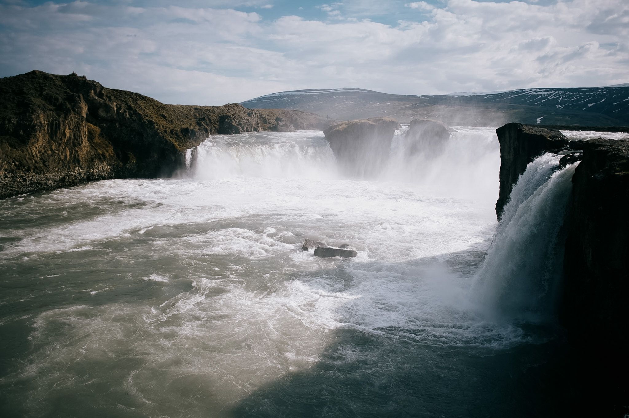 A wide horseshoe waterfall split by a rock in the middle, spray blowing up out of the pool