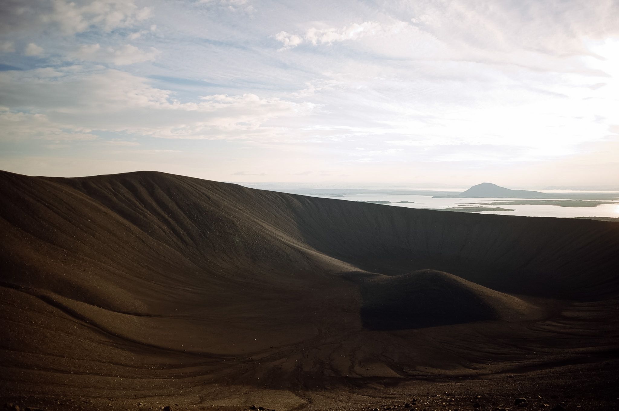 Looking down into a dark ash crater, its rim throwing a long shadow, a lake and a cone beyond